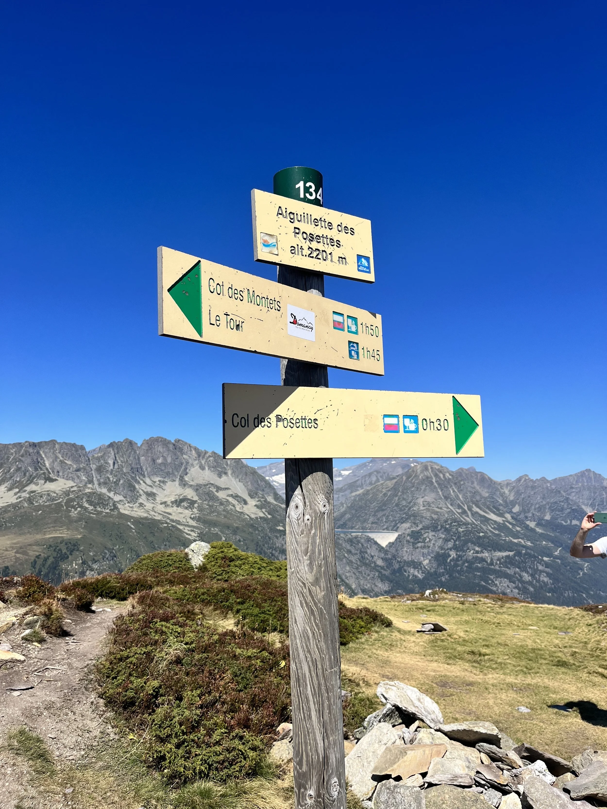 Trail signs on a wooden post in a mountain landscape with rocky terrain, green shrubs, and distant mountain peaks under a clear blue sky.