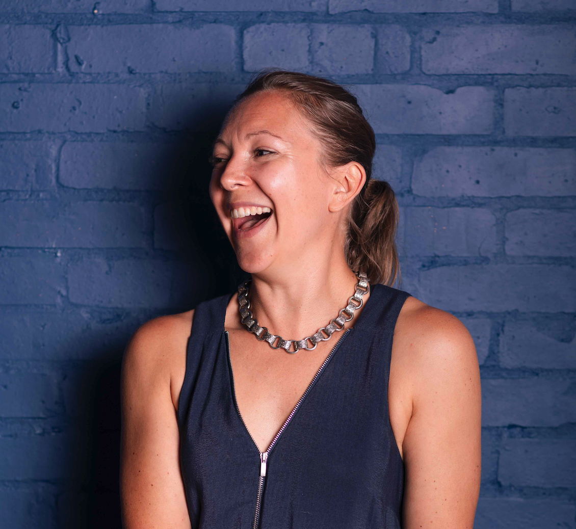 A woman with dark blonde hair styled in a ponytail, laughing and looking to the side, standing against a blue brick wall. She is wearing a navy sleeveless top with a zipper and a chunky silver necklace.