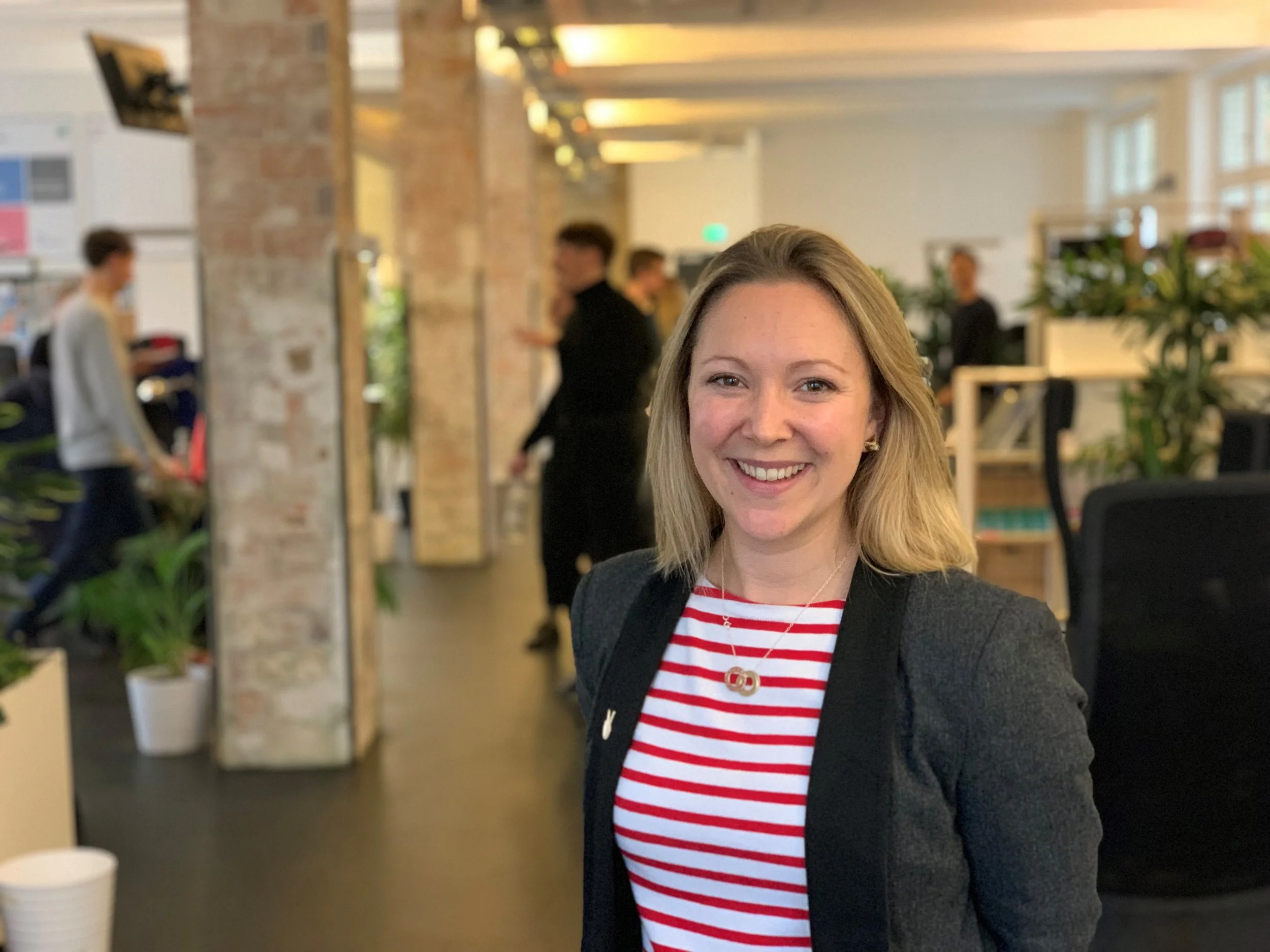 A woman smiling in an office setting, with people and plants in the background.