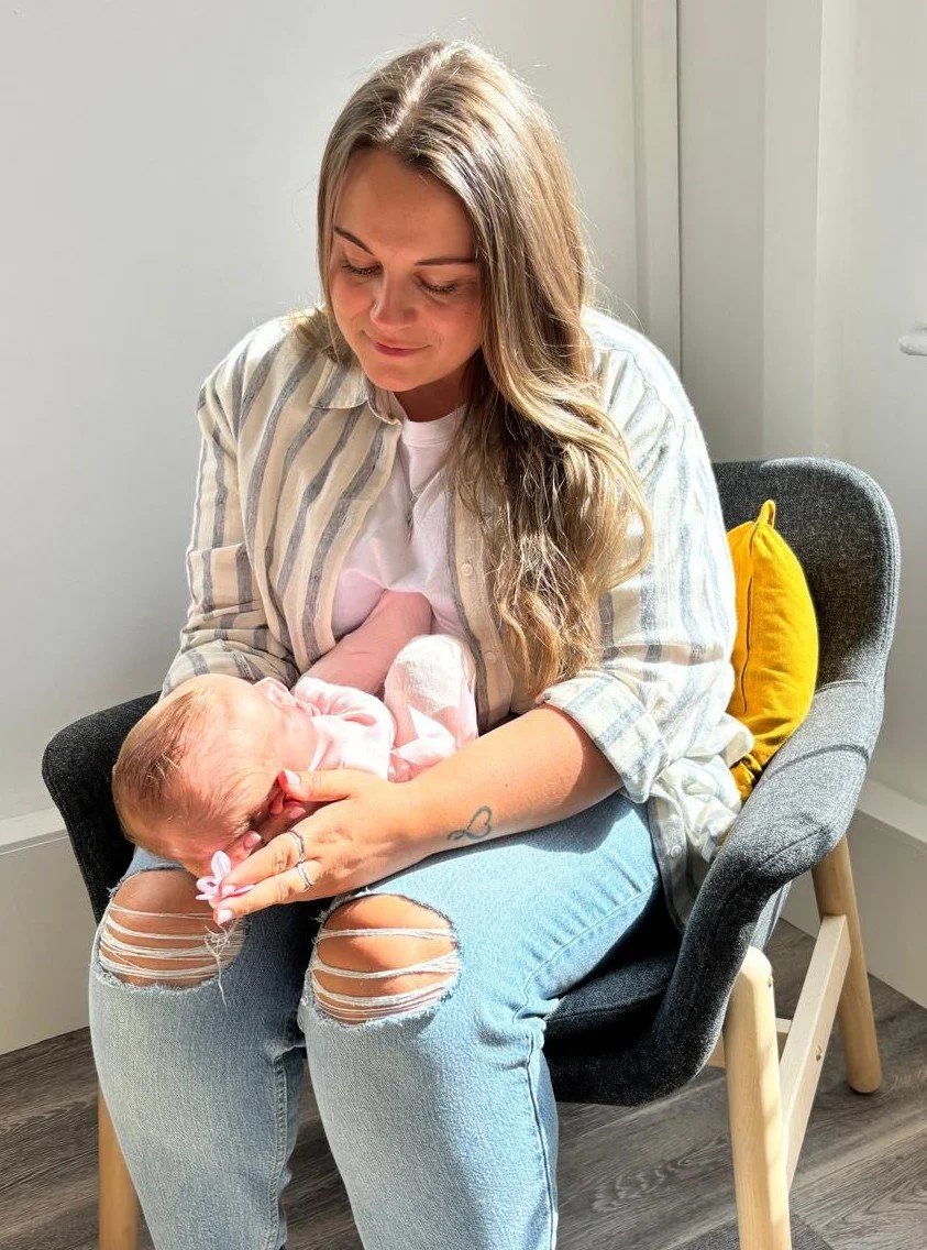 A woman sitting on a chair holding a newborn baby in her arms, looking down at the baby with a gentle expression.