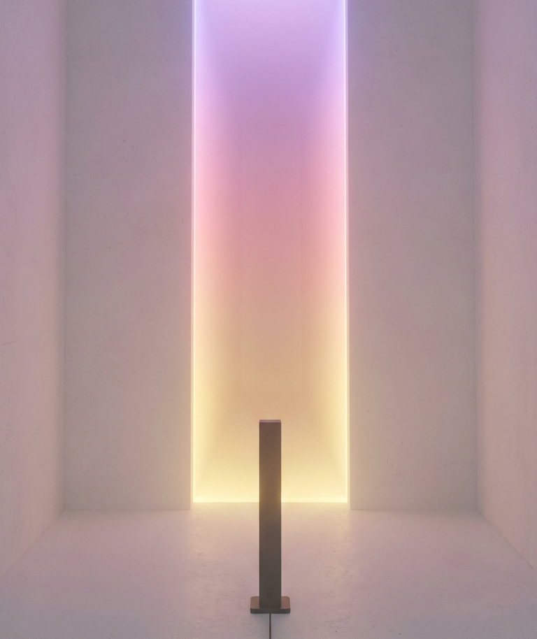 Empty room with a vertical, multicolored LED light panel on the wall and a small post in the center of the floor.