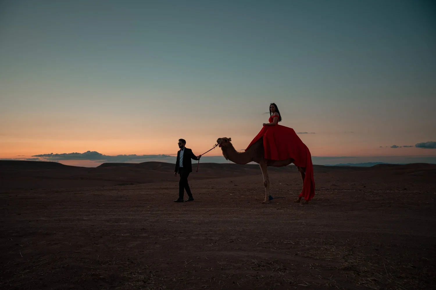A woman in a red dress sitting on a camel, being led by a man in a suit across a desert at sunset.