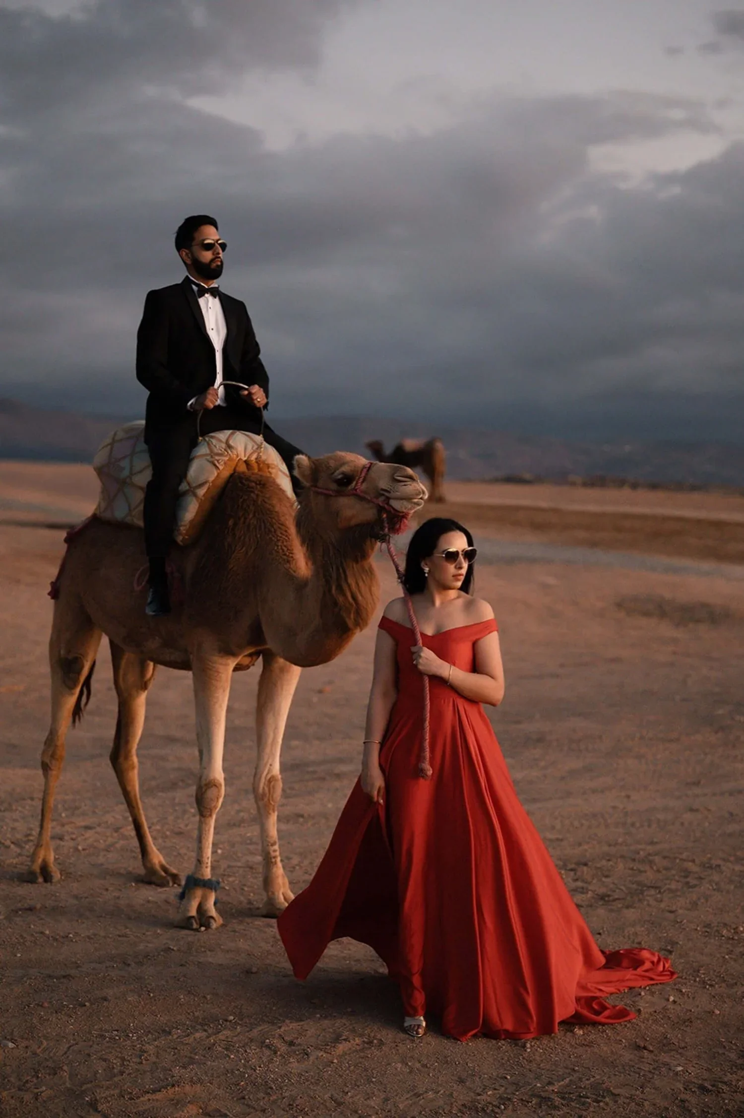 A man in a tuxedo riding a camel, a woman in a red gown holding the camel's leash, set against a desert landscape with a cloudy sky.