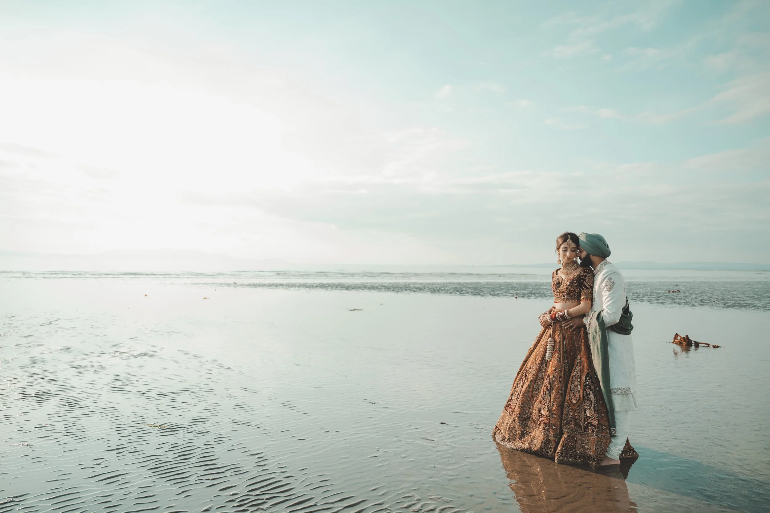 A couple dressed in traditional Indian wedding attire standing in shallow water at the beach, with the ocean and sky in the background.