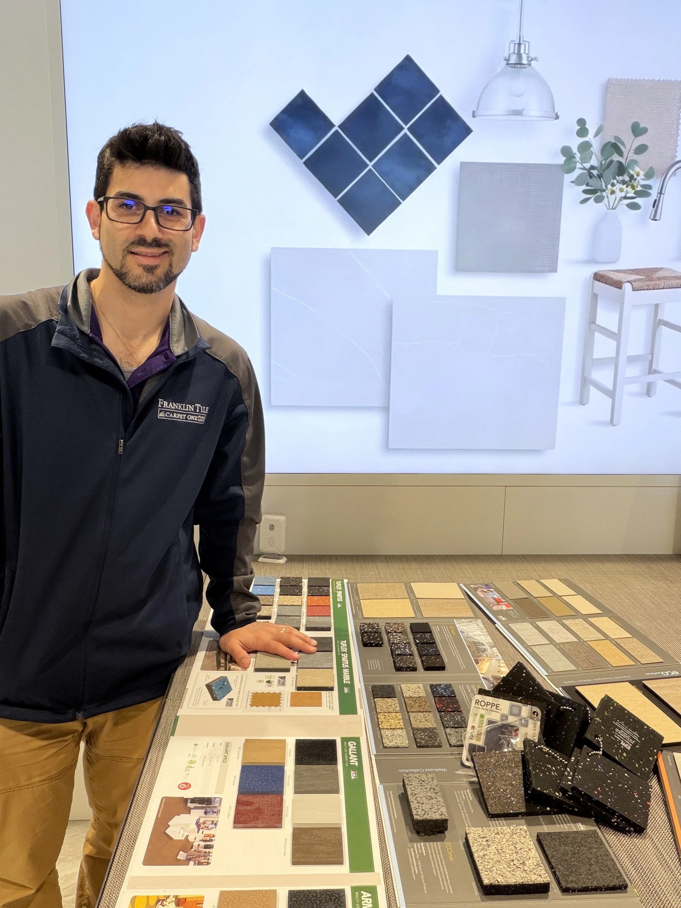 A man wearing glasses and a black and gray jacket stands by a display table of various flooring samples, including tiles, wood, and carpet options, in a showroom setting.