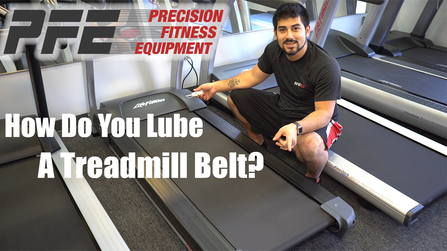 A man kneeling next to a treadmill, holding a lubricant spray, demonstrating how to lubricate a treadmill belt, in a gym with multiple treadmills and fitness equipment.