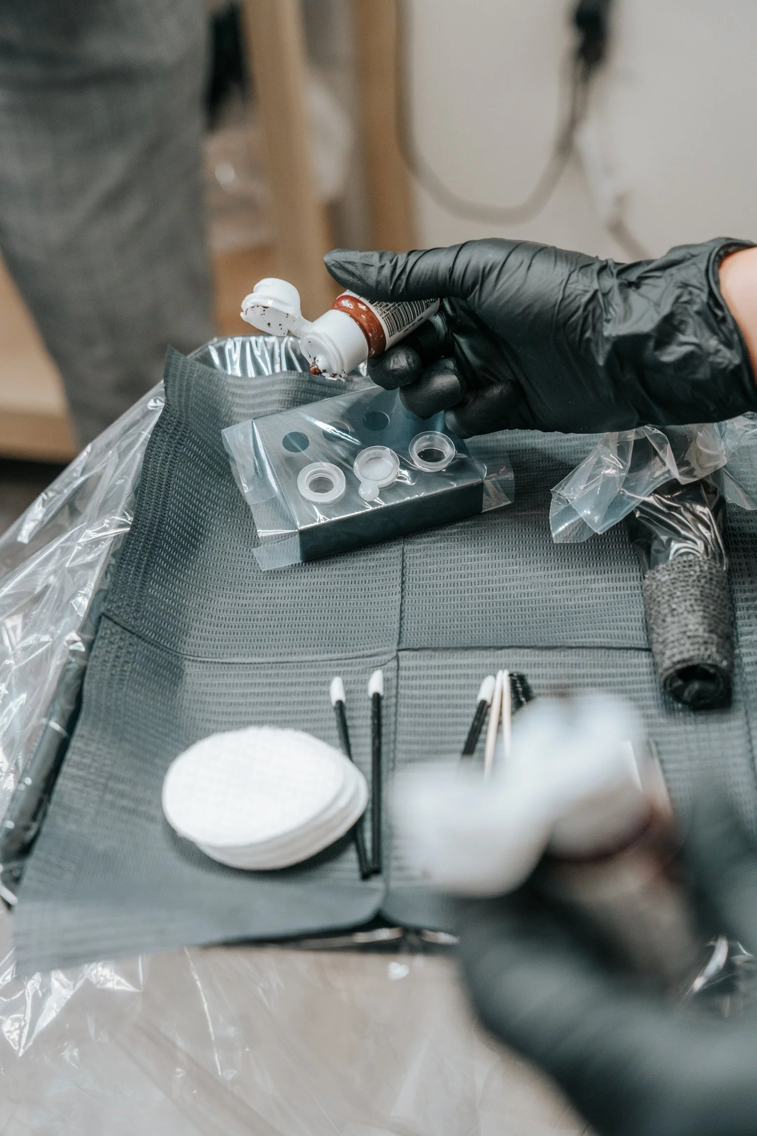 Close-up of a hand wearing a black glove handling a vial, surrounded by medical supplies including cotton swabs, small containers, and medical tools, on a gray tray.