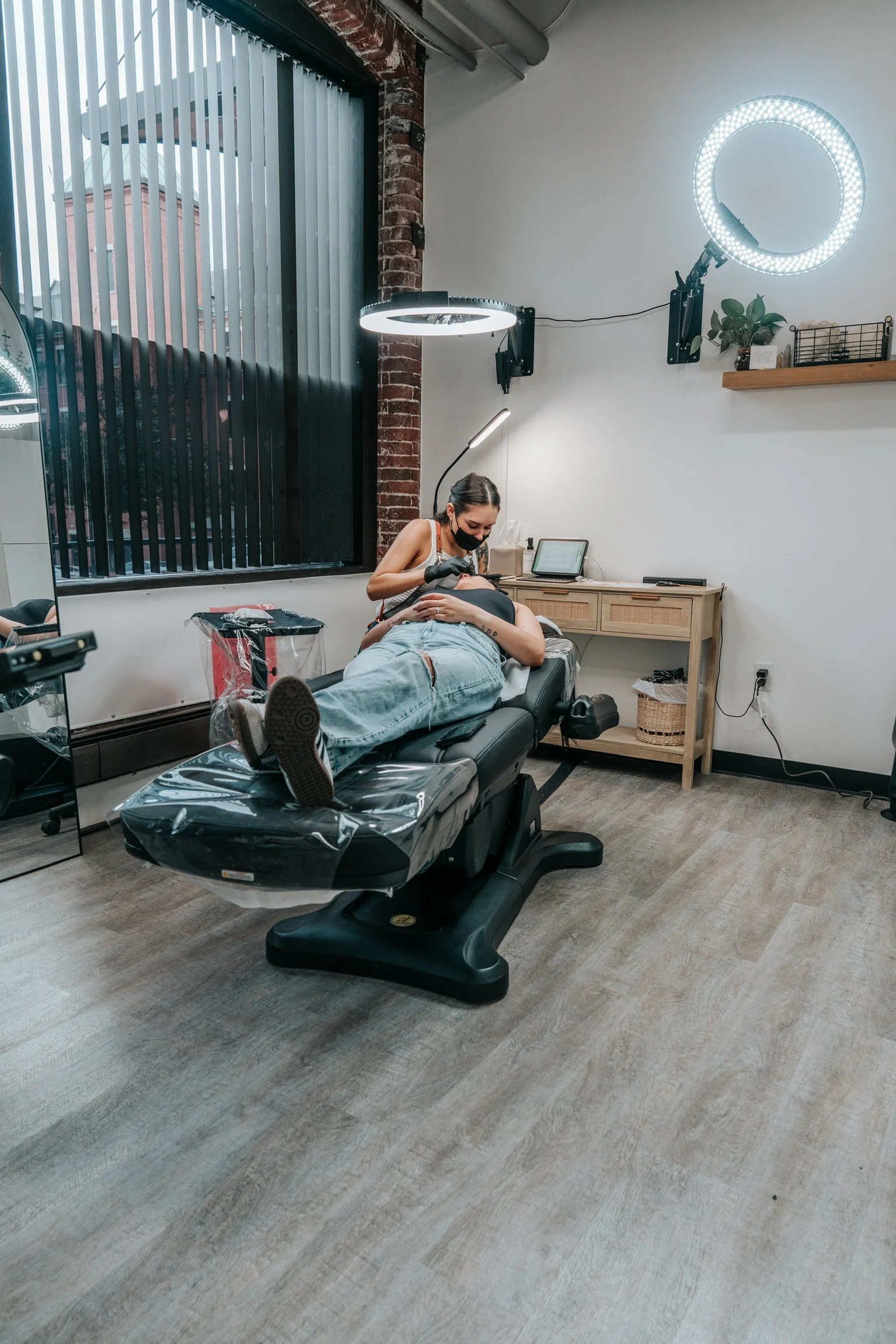 A woman receiving a cosmetic tattoo procedure from a tattoo artist in a modern tattoo studio with a ring light and a large window.