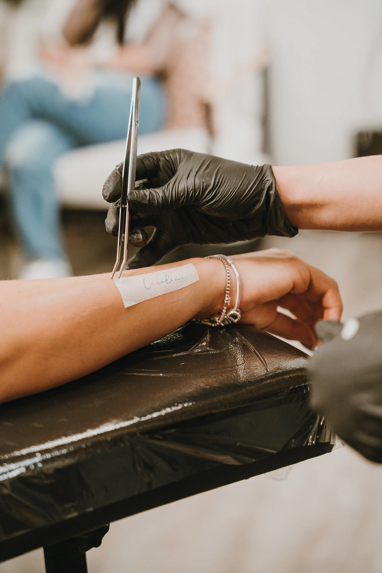 A person getting a tattoo on their forearm from an artist wearing black gloves. The tattoo artist is using tweezers, and the person's forearm is labeled with a tape that says 'Caribbean.' The person is wearing bracelets, and there is a person sitting