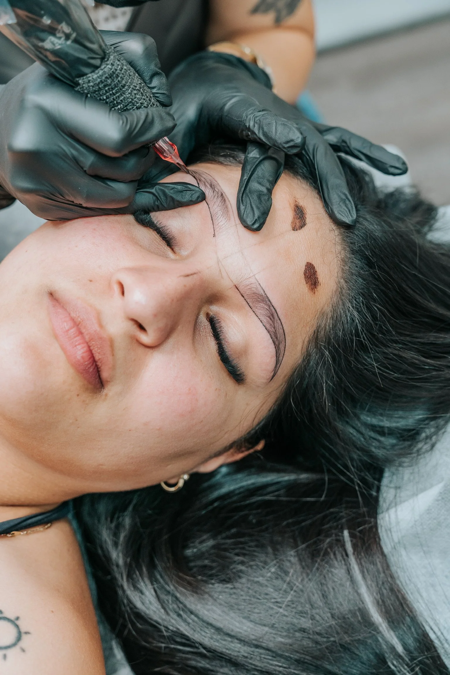 A woman lying down with her eyes closed receives a cosmetic tattoo procedure on her eyebrow from a professional wearing black gloves.