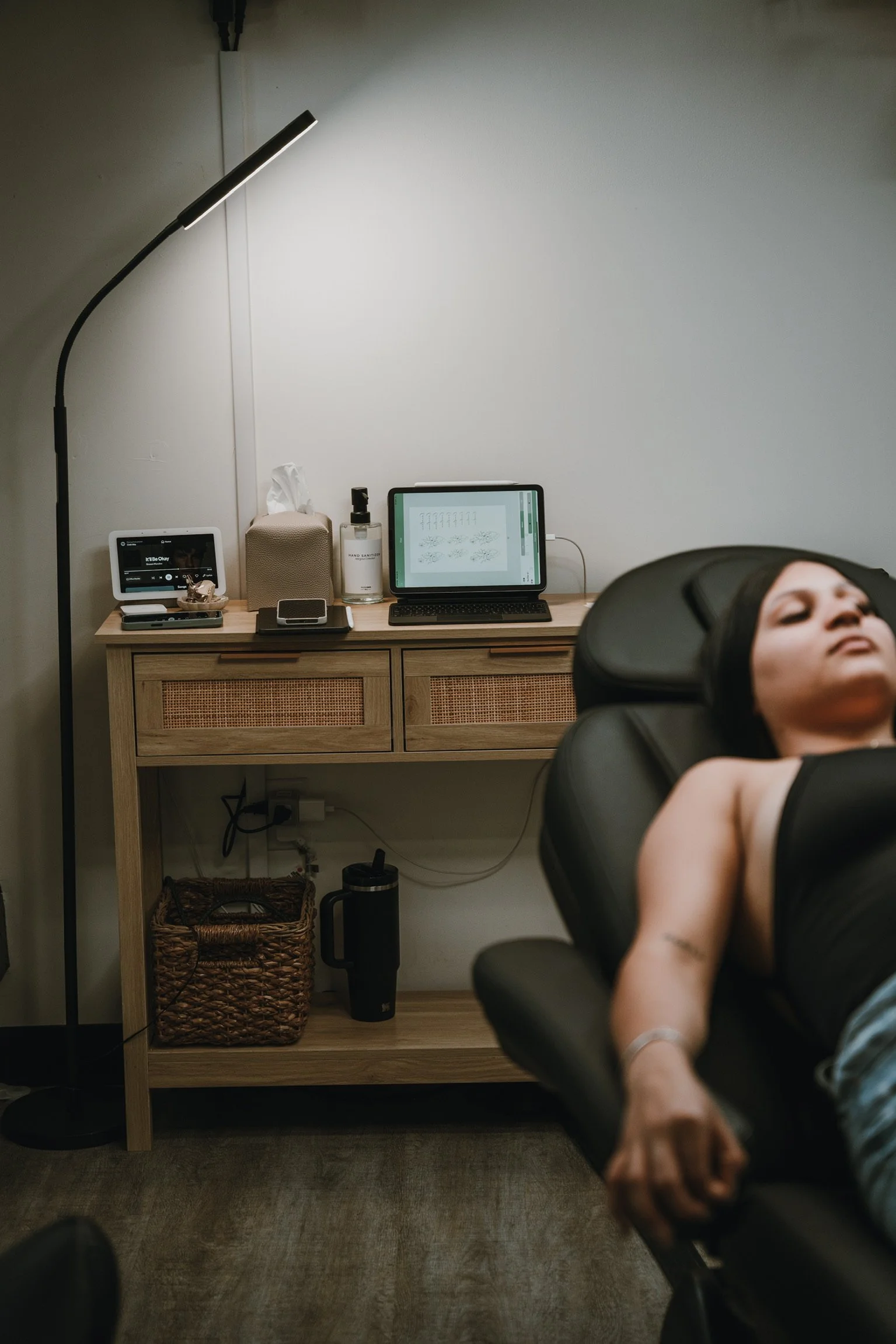 A woman lying on a black reclining chair, with her head tilted back, in a room with a wooden desk and various electronic devices, including a computer and a tablet.
