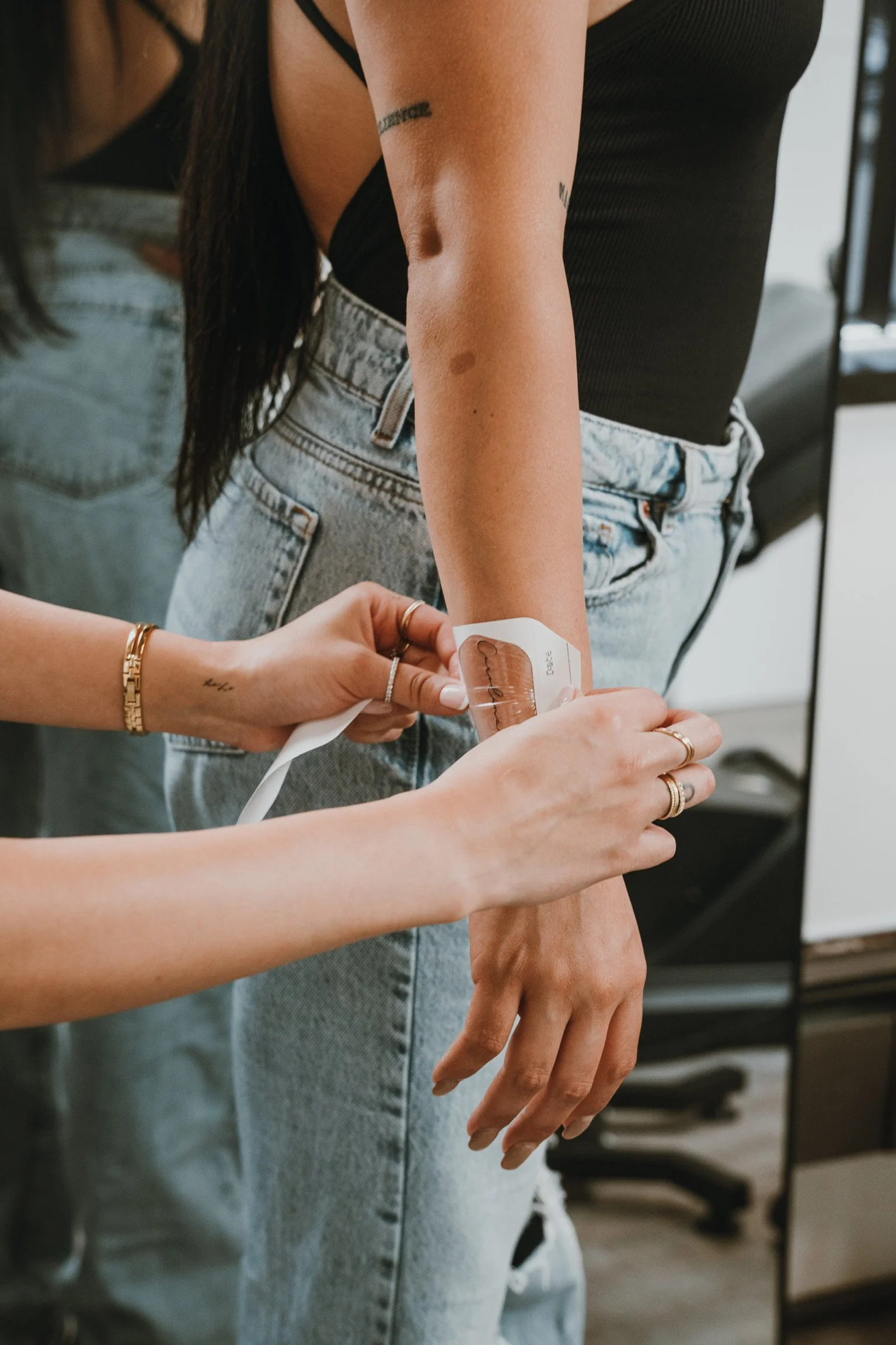 A person is having a temporary tattoo applied to their forearm by another individual. The person receiving the tattoo is wearing a black sleeveless top and light blue distressed jeans. The person applying the tattoo has bracelets and rings on their f