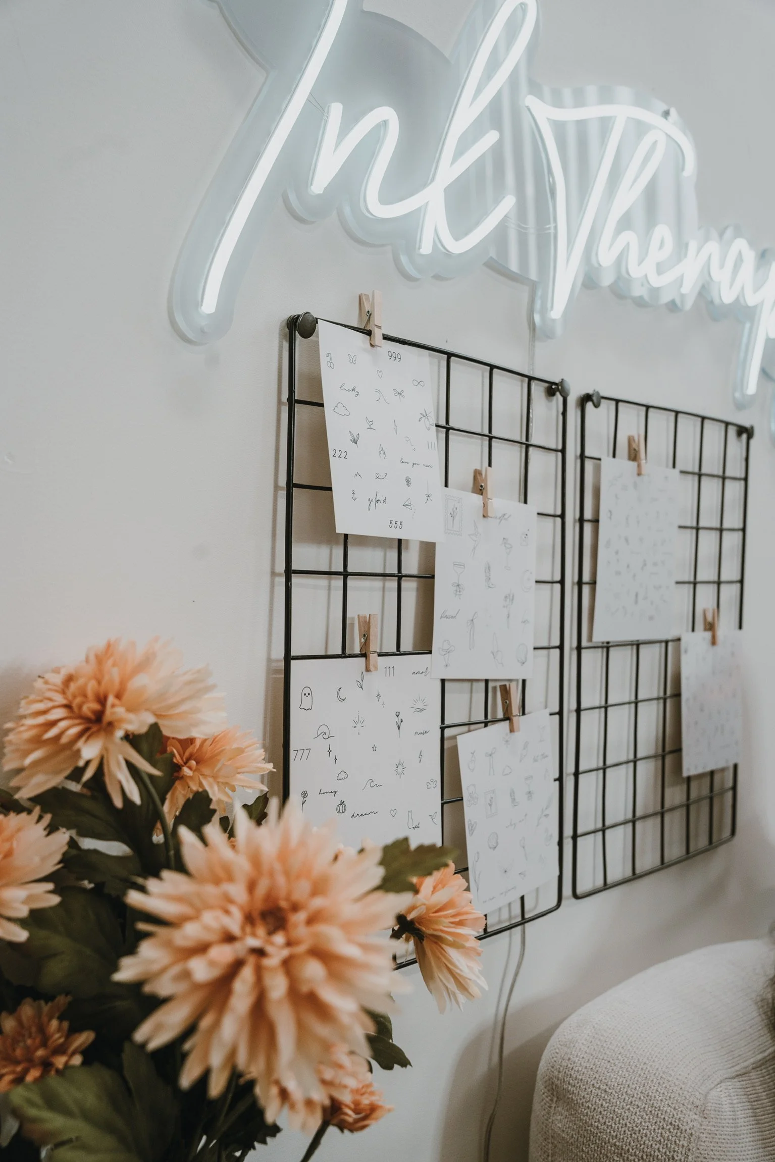 Decorative wall with a neon sign that says 'thank you', and two black wire grid panels with white paper sheets attached by wooden clothespins, along with a bouquet of peach-colored flowers in the foreground.