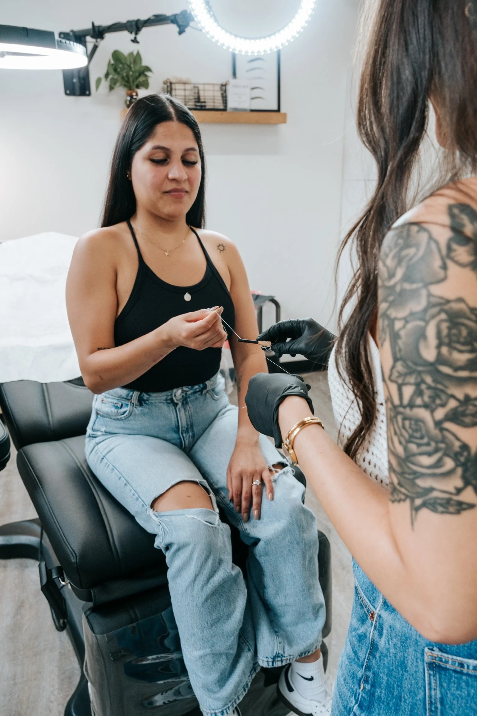 A woman sitting on a black chair receiving a tattoo or piercing from a person wearing black gloves in a tattoo studio.