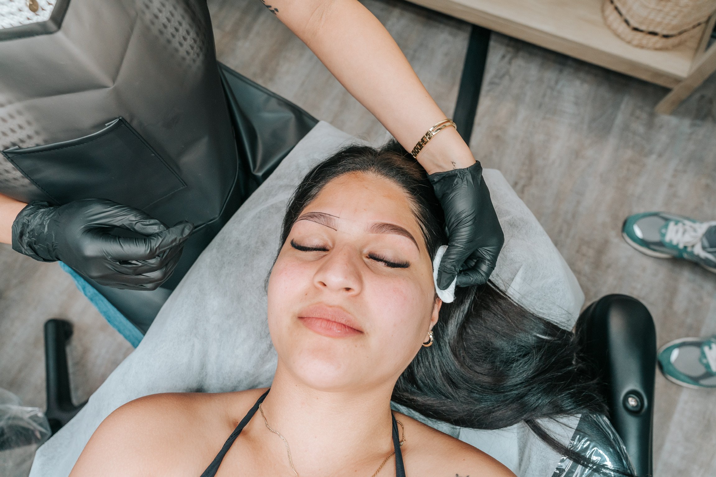 A woman is lying down with her eyes closed, getting a facial treatment from a professional wearing black gloves.