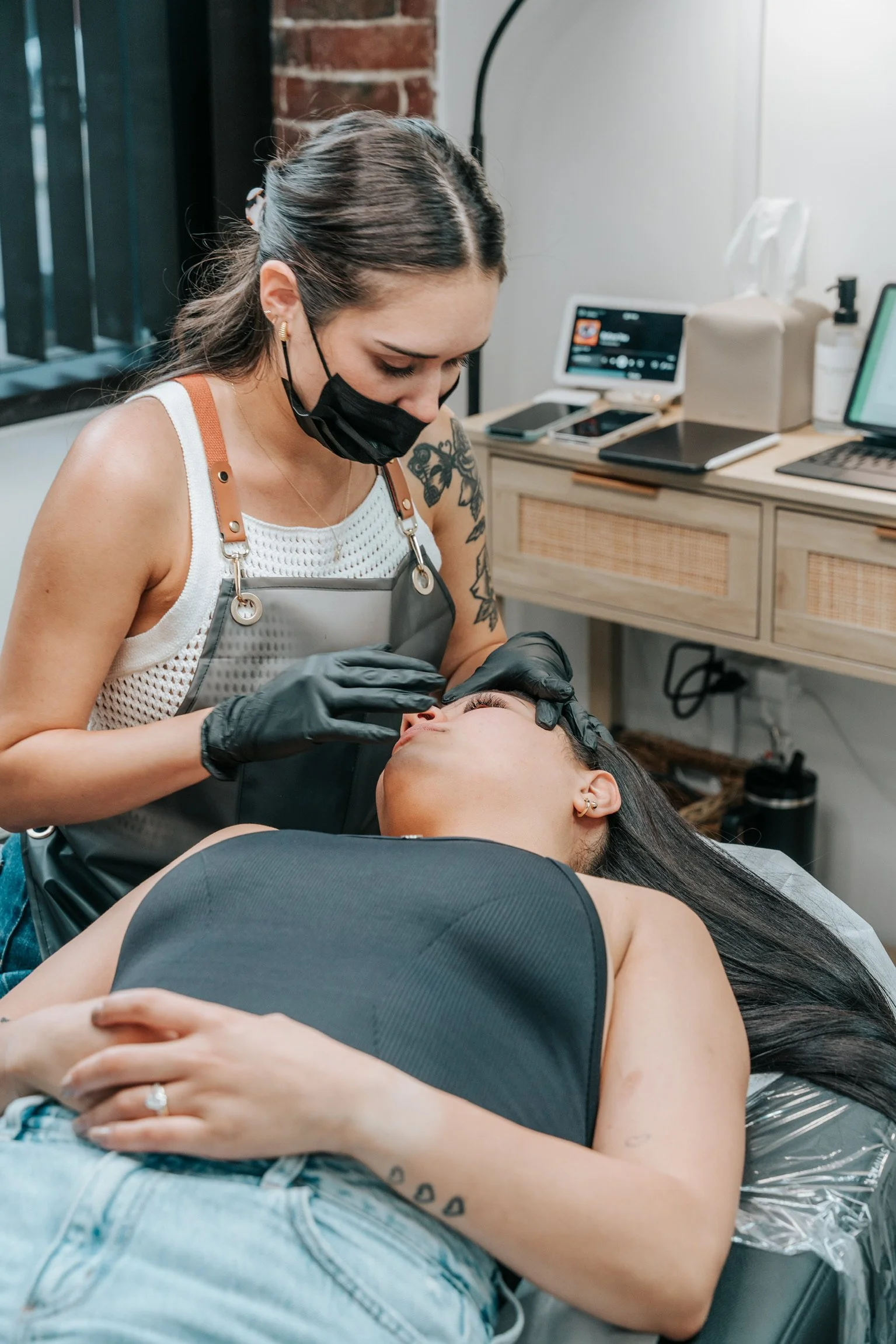 Tattooed woman receiving eyelash extension treatment from a technician in a cosmetic clinic.