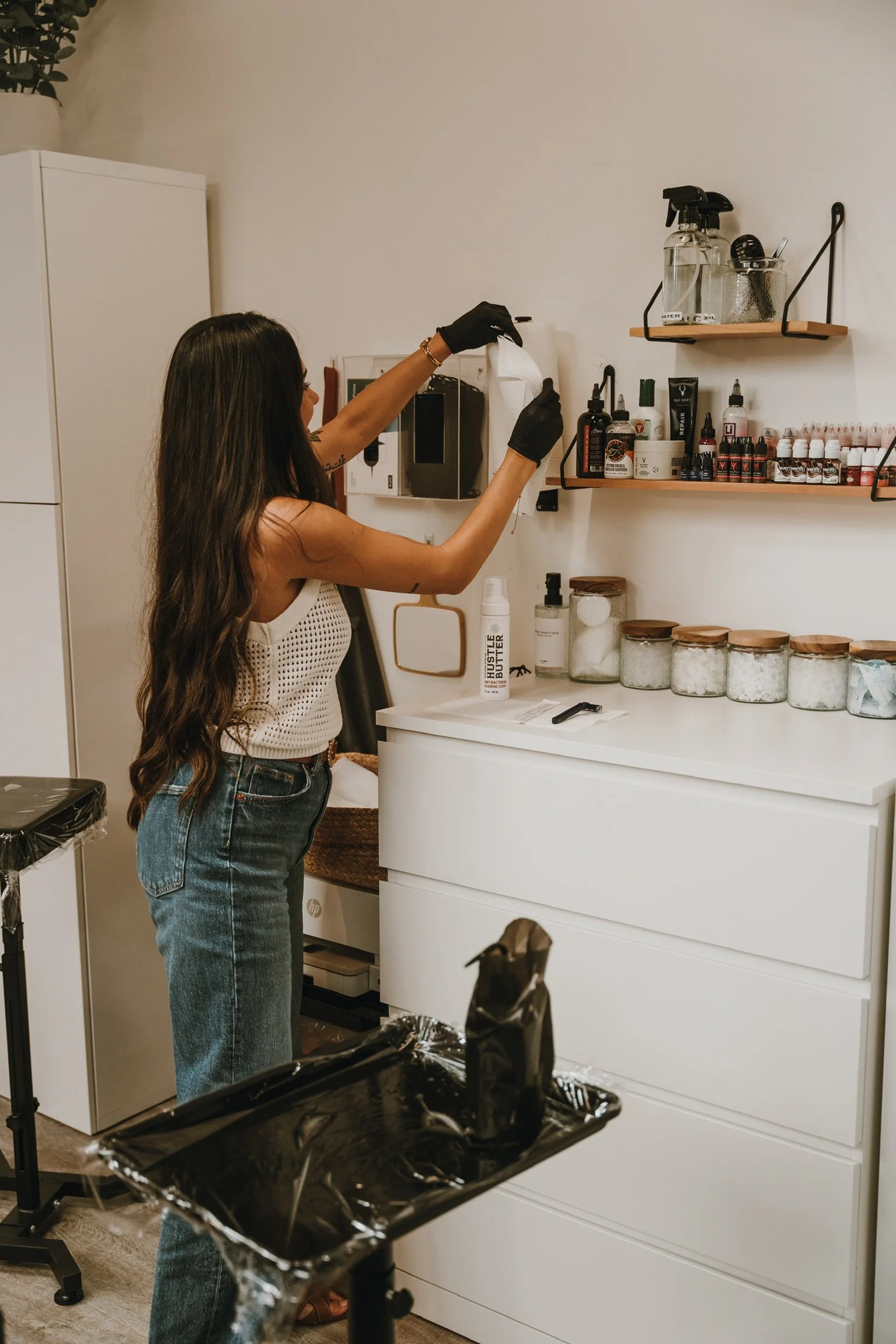 A woman with long, dark hair wearing a white sleeveless top, high-waisted jeans, and black gloves is working in a beauty or tattoo studio. She appears to be preparing equipment or supplies, with shelves of small bottles, jars, and equipment behind he