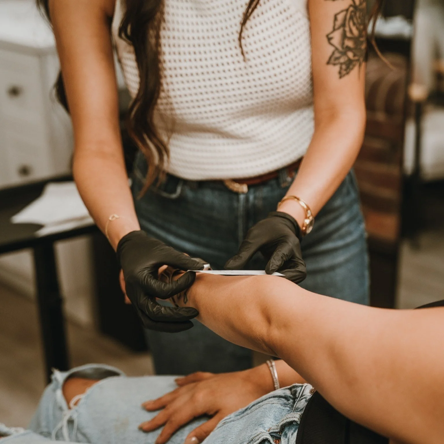 A person wearing a white, mesh sleeveless top and jeans receiving a tattoo on their arm from a tattoo artist. The artist is wearing black gloves and holding a tattoo gun. The tattoo artist has a floral tattoo on their upper arm.