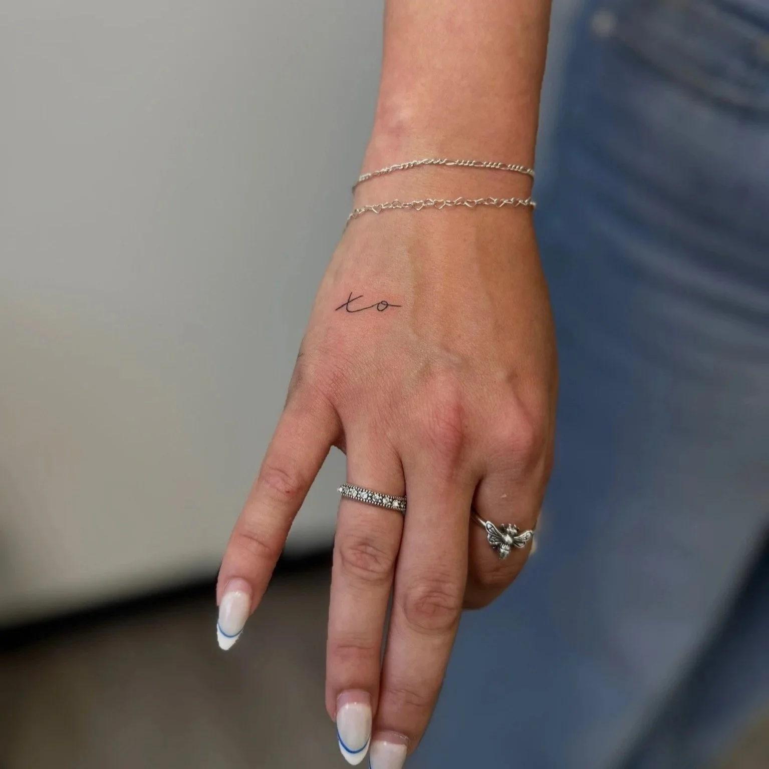 Close-up of a woman's hand with a small tattoo reading 'xo' on the wrist, wearing two silver rings, one with diamonds, and a silver bracelet, against a background of a blue shirt and neutral wall.