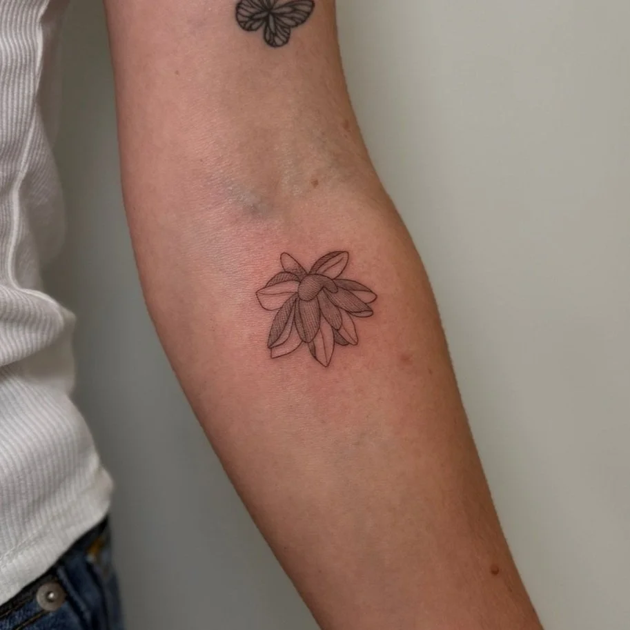 A close-up of a person's inner arm showing a freshly done outline of a flower tattoo with petals, and part of another tattoo of a butterfly above it.