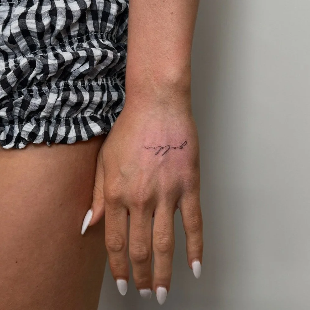 A person's right hand resting against their thigh, showing a delicate tattoo that says 'dream' on the wrist, with painted white nails. The person is wearing black and white checkered shorts and standing against a plain wall.