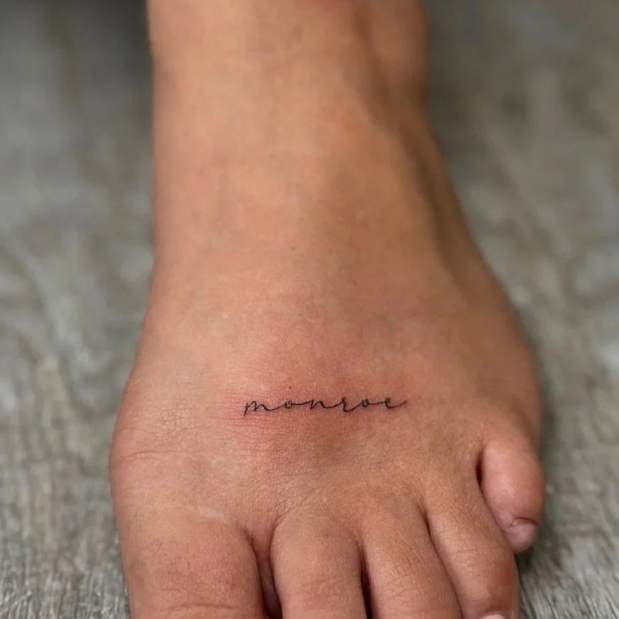 A small, black tattoo of the word "monroe" in cursive on the top of a person's foot.