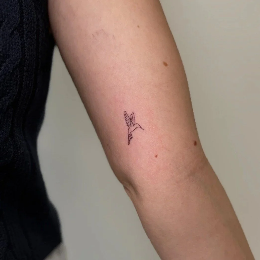 A person's arm with a small, minimalist tattoo of a hummingbird in flight.