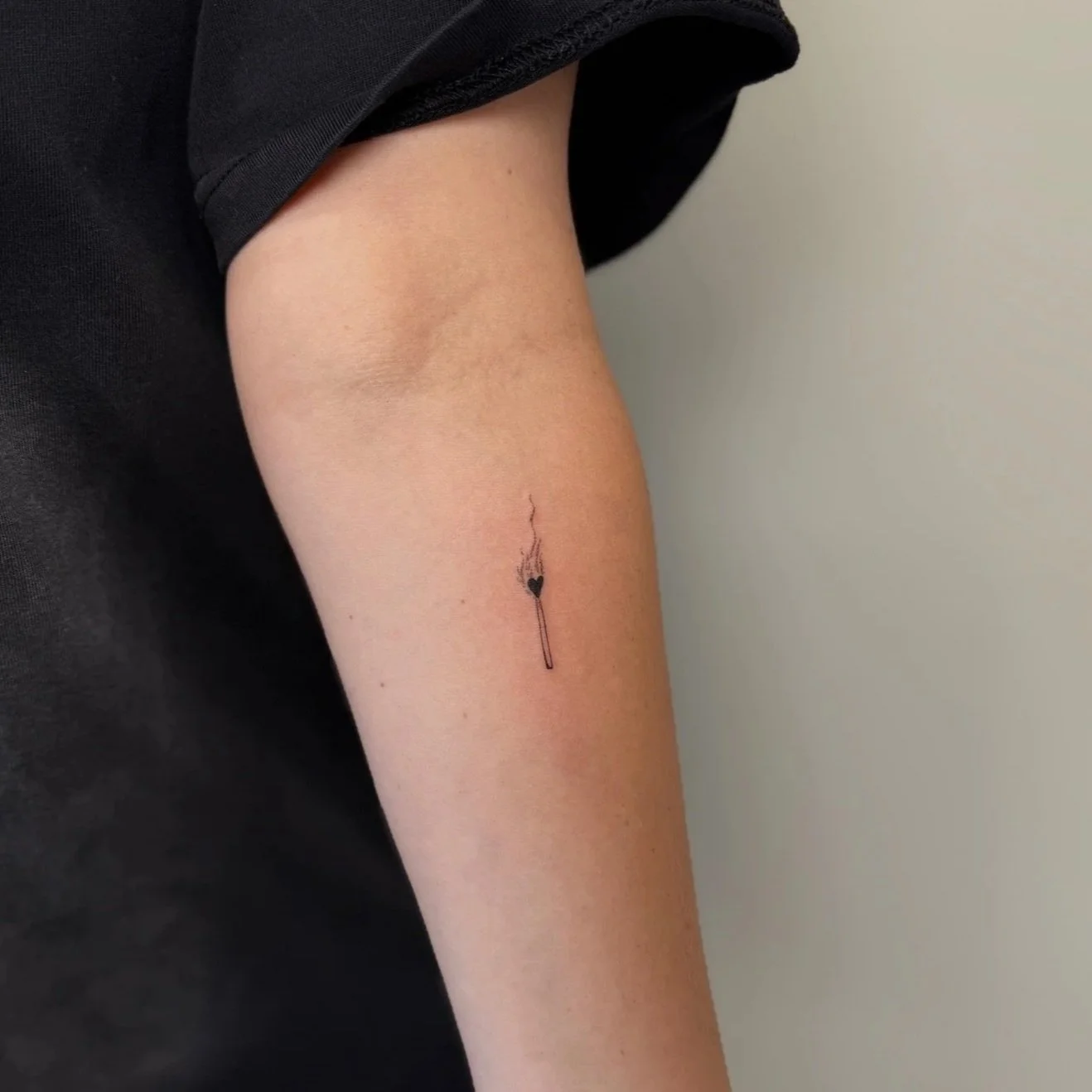 A tattoo of a burning matchstick with a heart-shaped flame on a person's upper arm.