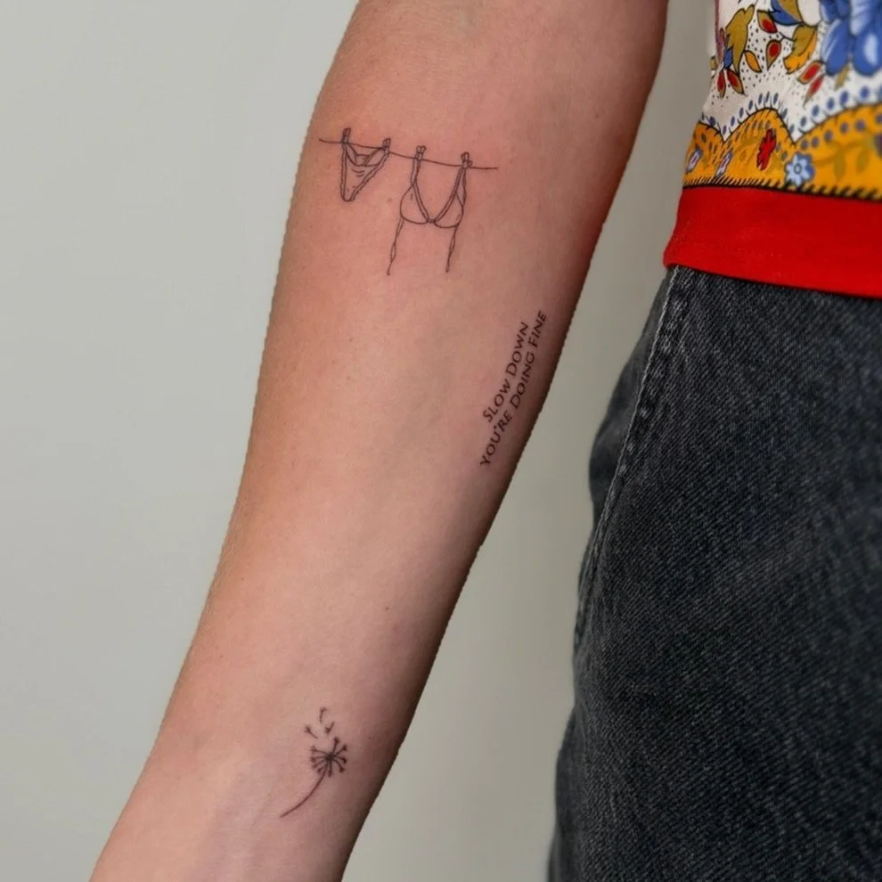 Tattoo of a clothesline with underwear and a bra, a quote reading "Slow down, you're doing fine," and a dandelion seed head blowing in the wind.