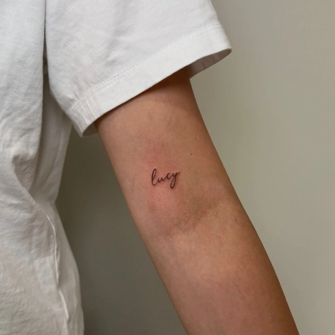 Person's upper arm with a small, black, cursive tattoo of the word 'lucy' on the inner side, next to a slightly discolored patch of skin, wearing a white t-shirt.