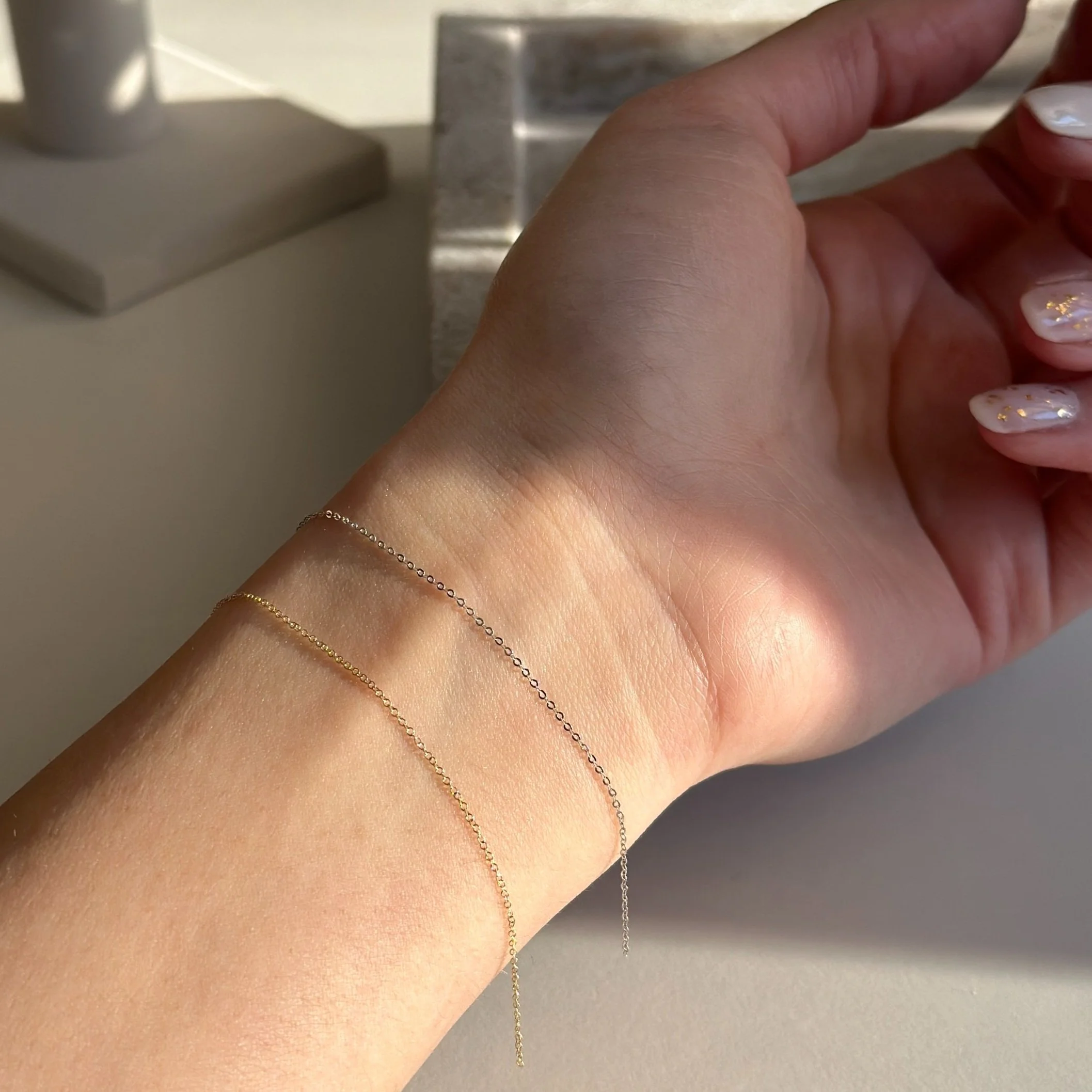 A person's wrist with three delicate gold and silver chain bracelets resting on a beige surface, with sunlight casting shadows.