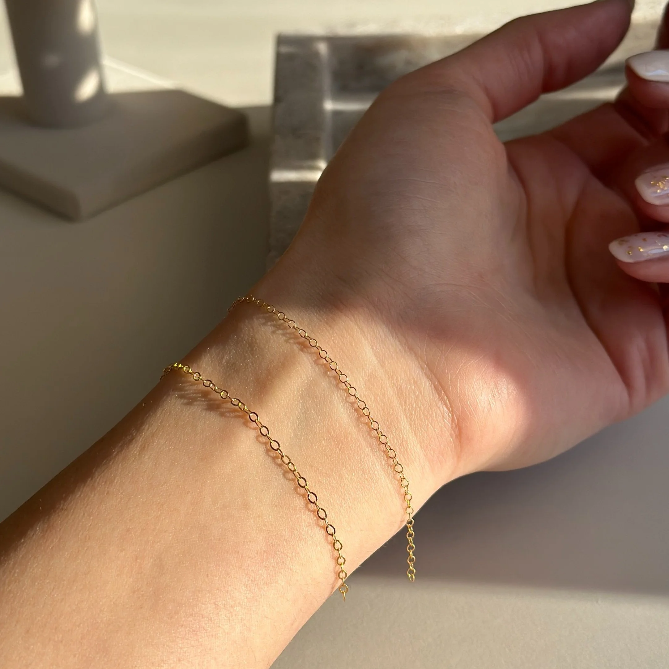 A person's wrist with two delicate gold chain bracelets, one slightly longer than the other, against a neutral background.