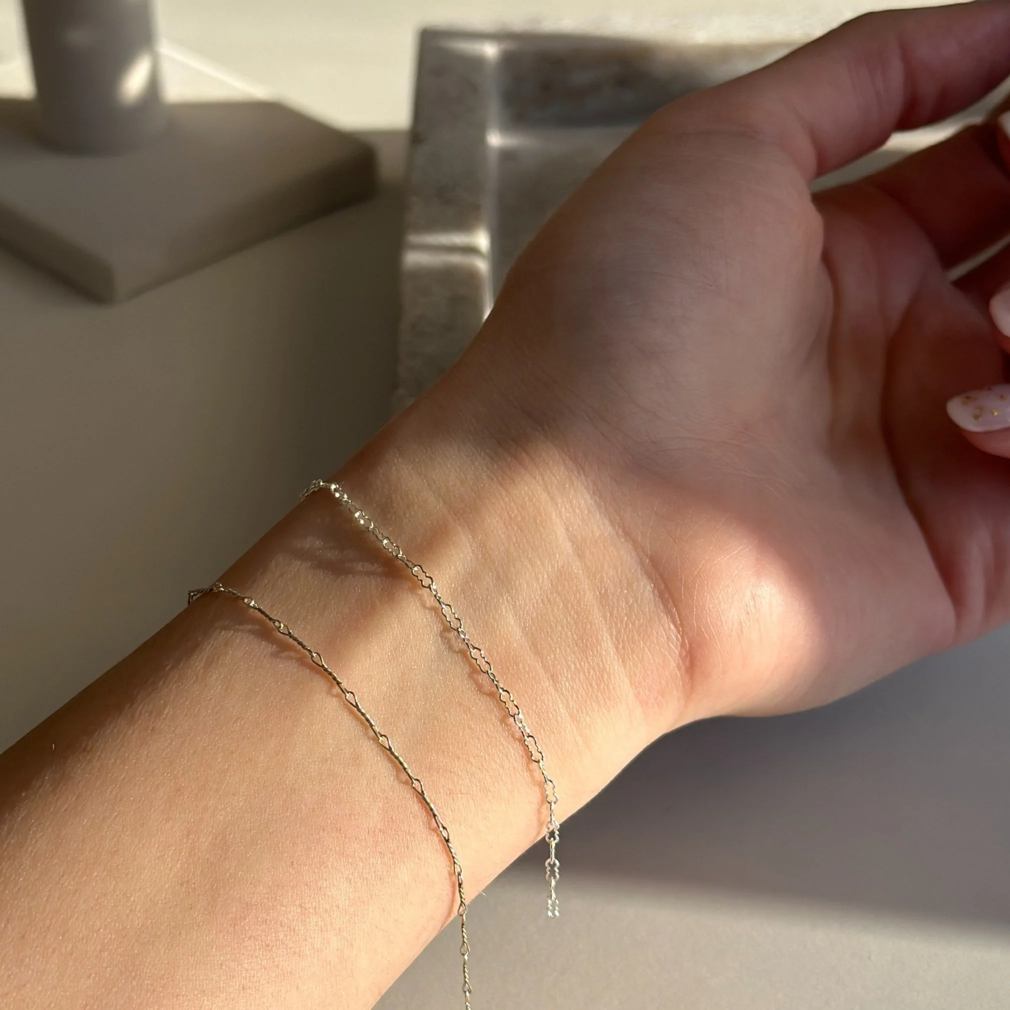 A person's wrist with two delicate silver chain bracelets, resting on a flat surface near a lamp and metallic object.
