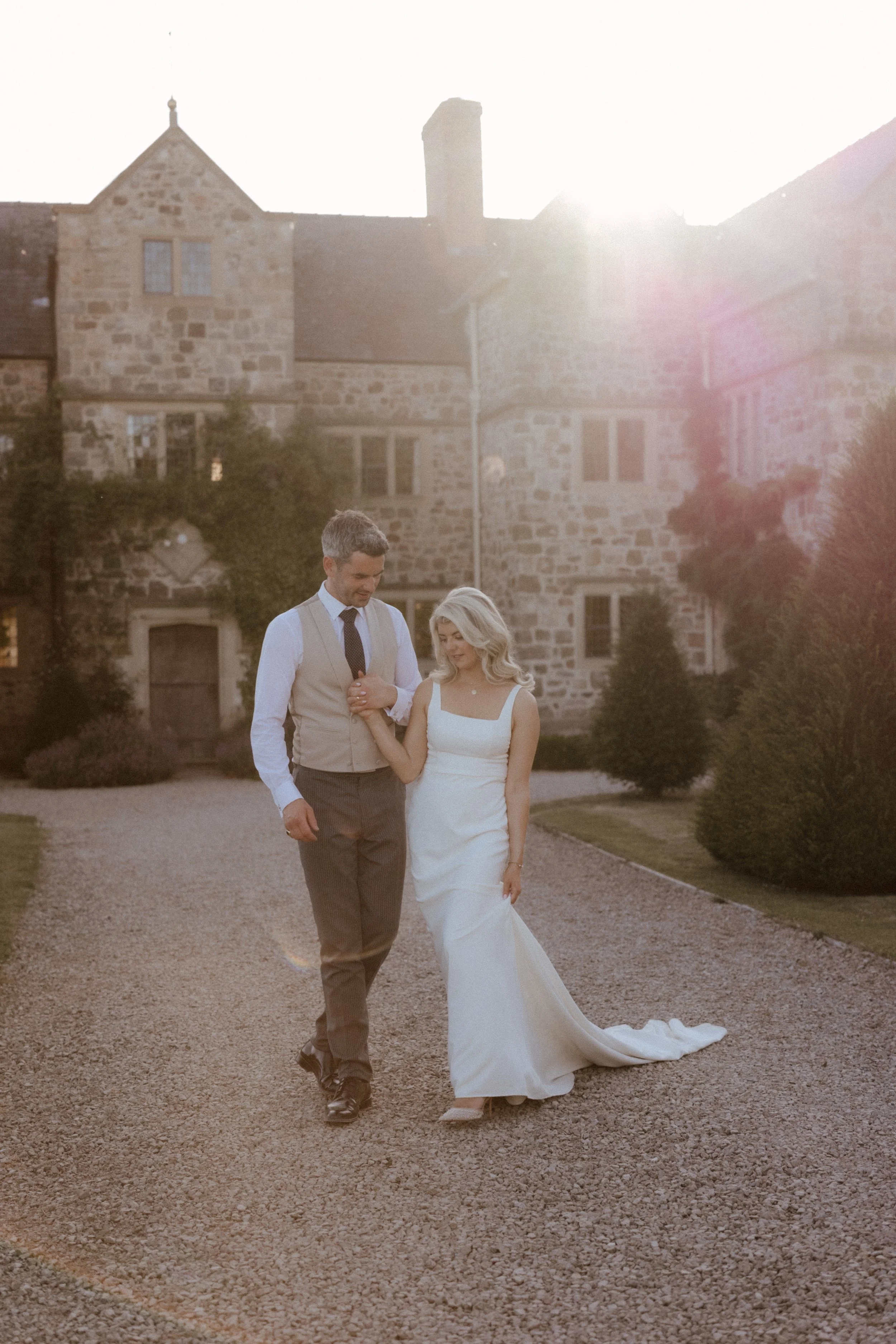 A couple walking hand in hand on a gravel path outside a stone mansion during sunset, with the bride in a white wedding dress and the groom in a beige vest and tie.