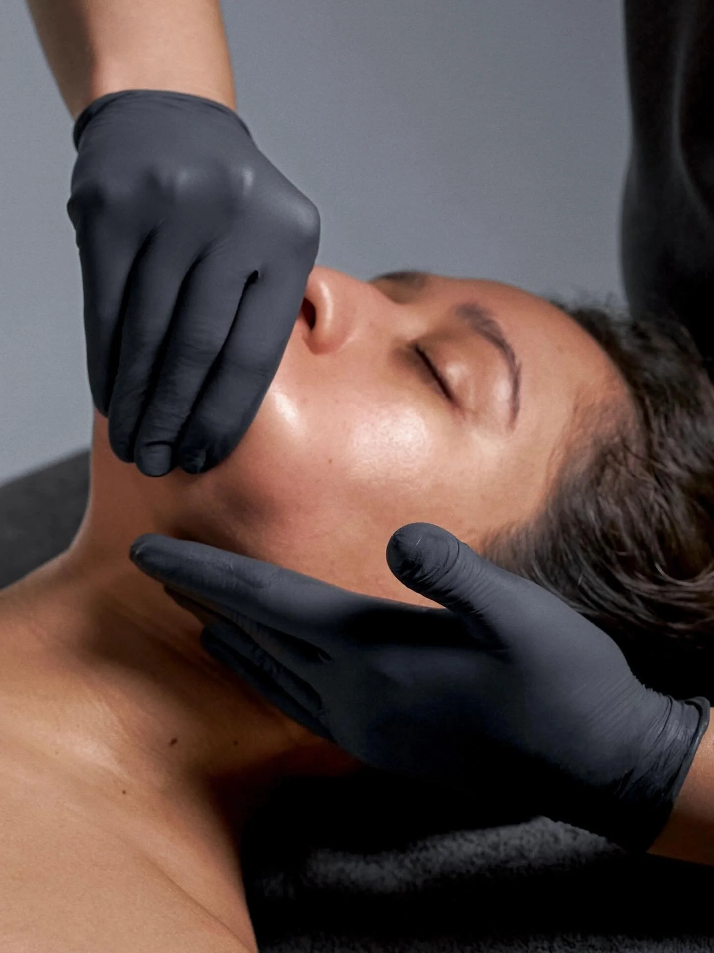 A person receiving a facial massage or skincare treatment while lying down, with hands in black gloves gently touching their face and neck.
