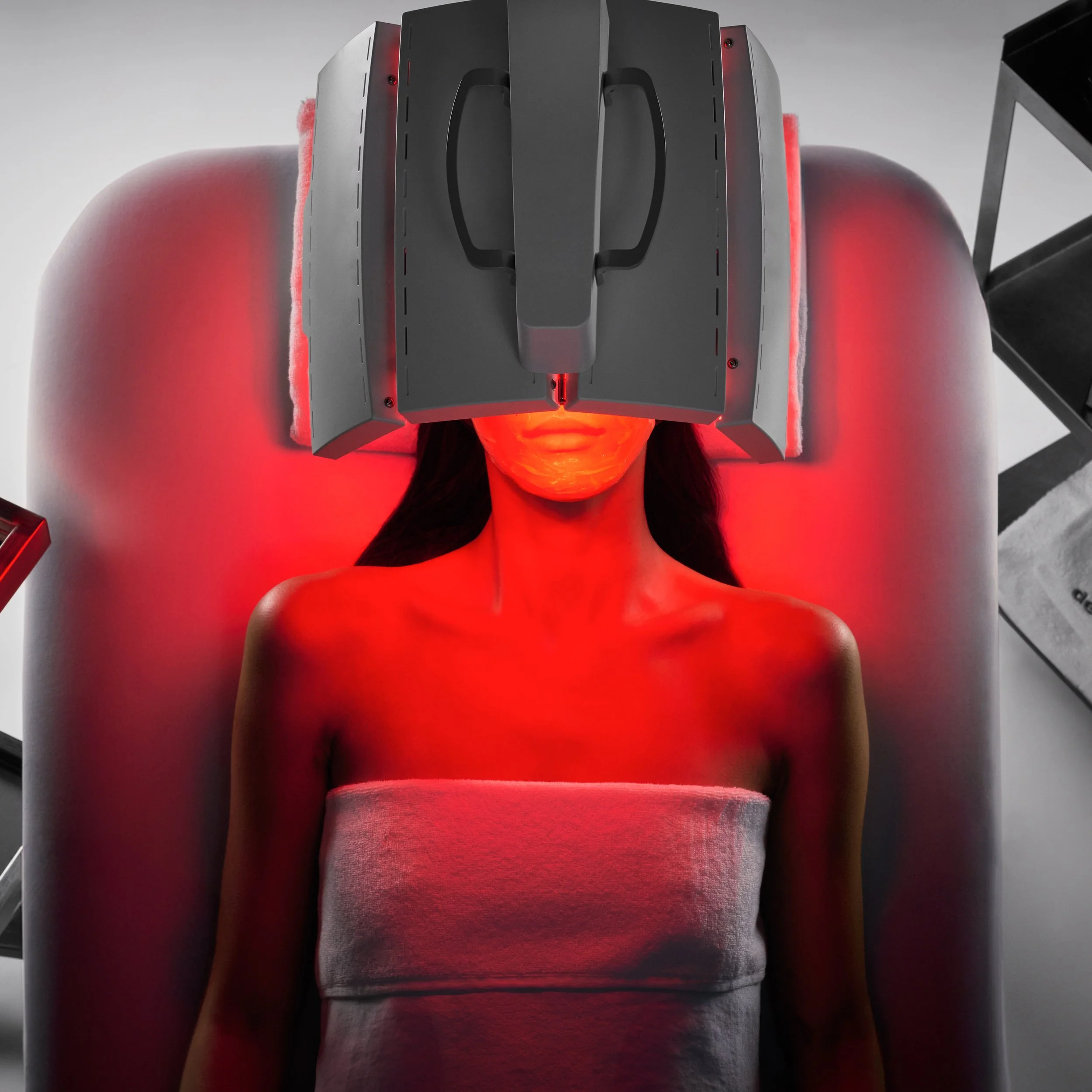 Person using a futuristic virtual reality headset emitting red light, lying on a bench, with a gray background.