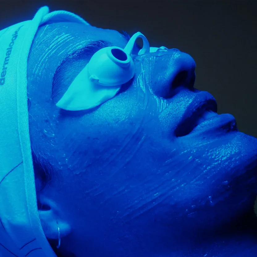 Close-up of a person's face illuminated with blue light, with a white medical device resting on their closed eyelids, possibly during a medical or skincare procedure.