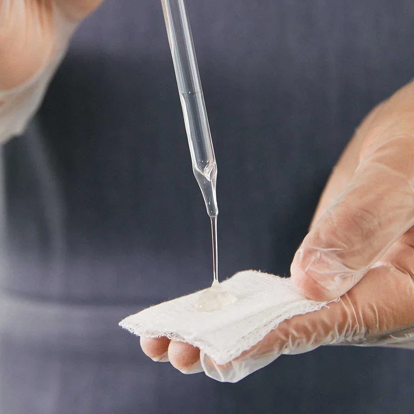 A person wearing disposable gloves using a dropper to extract or put a clear liquid onto a piece of white gauze or cloth.