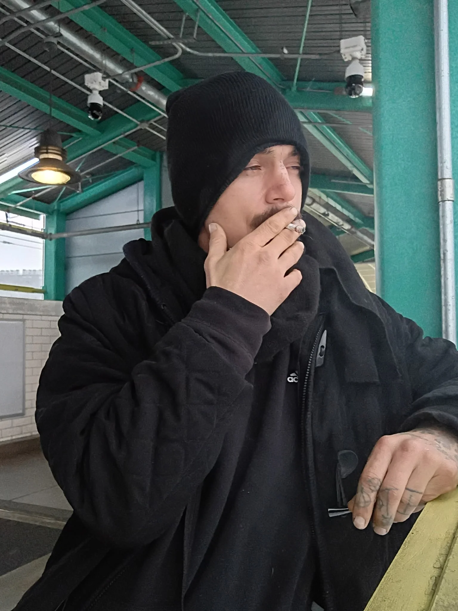 A man wearing a black beanie and black jacket is sitting and smoking a cigarette at an outdoor seating area, with a green and metal structure overhead.