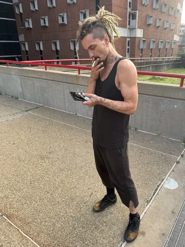 Young man with dreadlocks and tattoos standing outdoors, looking at his smartphone, wearing a black tank top, black pants, and boots, in an urban setting.