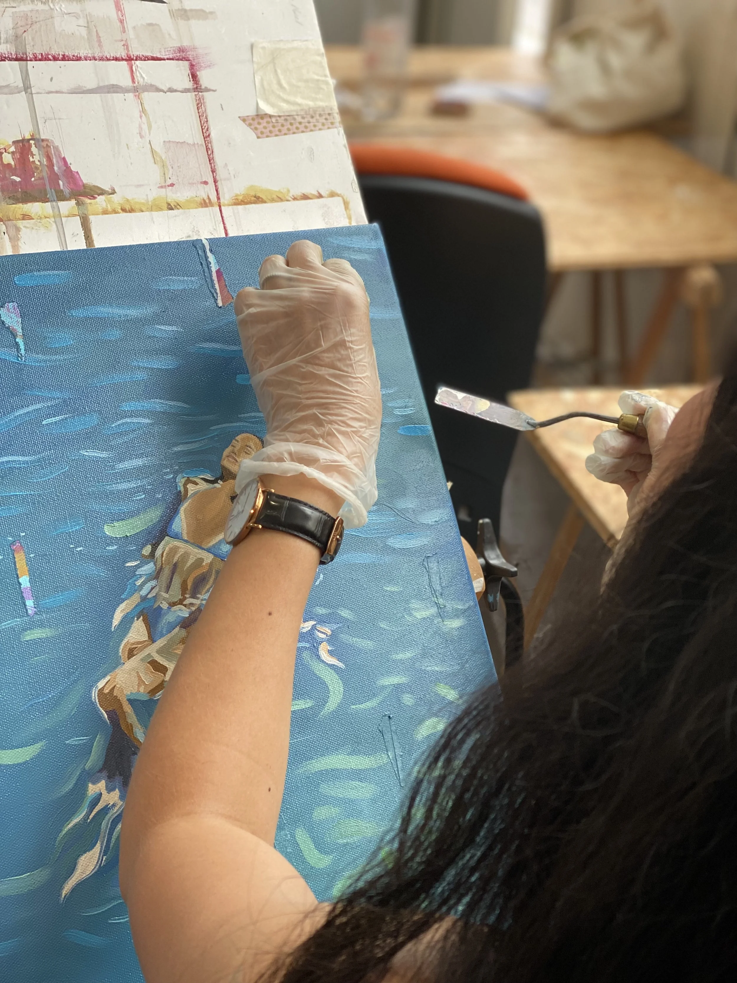 Une personne peignant une scène maritime avec un pinceau, portant une montre noire et un gant en plastique, sur une toile posée sur un chevalet.