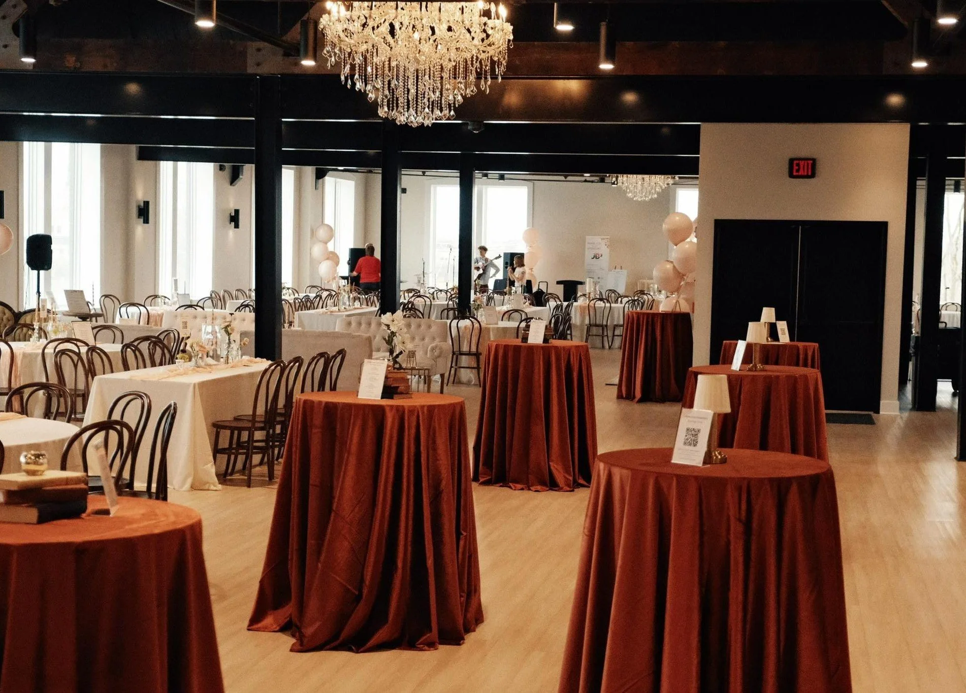 A spacious event hall decorated with chandeliers, round tables covered with brown and white tablecloths, chairs arranged around the tables, and a few people setting up or preparing for an event.