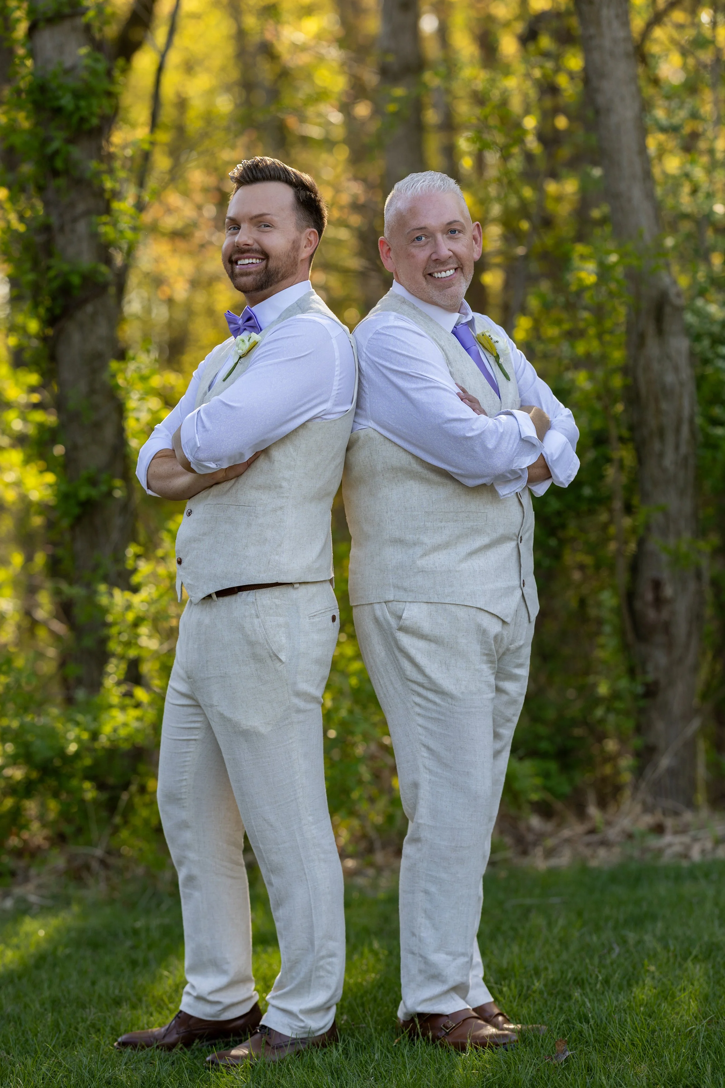 Two men standing outdoors back to back with arms crossed, smiling, dressed in light-colored suits with purple accents, in a wooded area with green grass and trees.