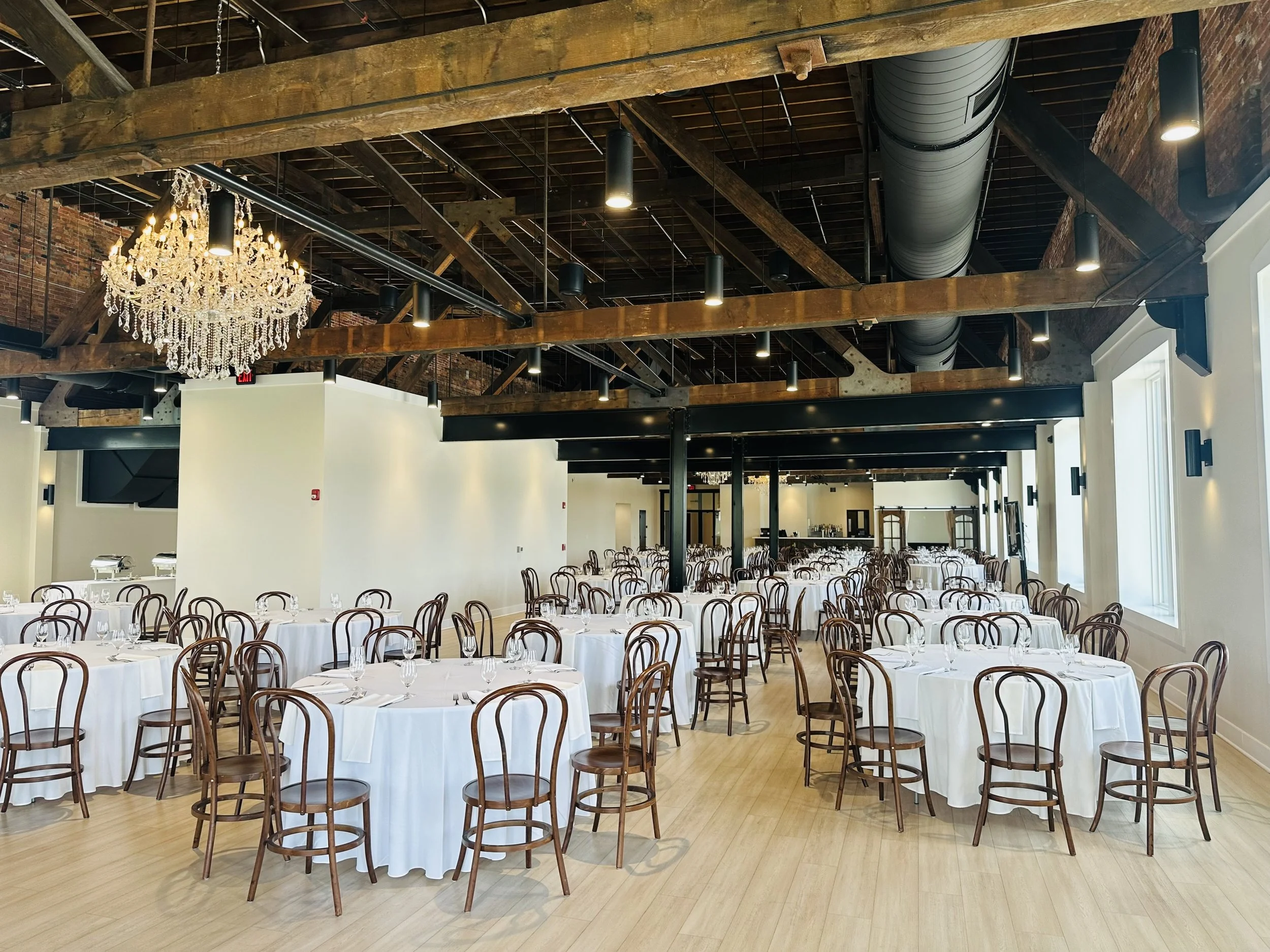 A spacious event hall set up for a formal gathering with round tables covered in white tablecloths and brown wooden chairs. The hall features an exposed wooden beam ceiling with industrial-style ductwork and black lighting fixtures. A large, ornate c