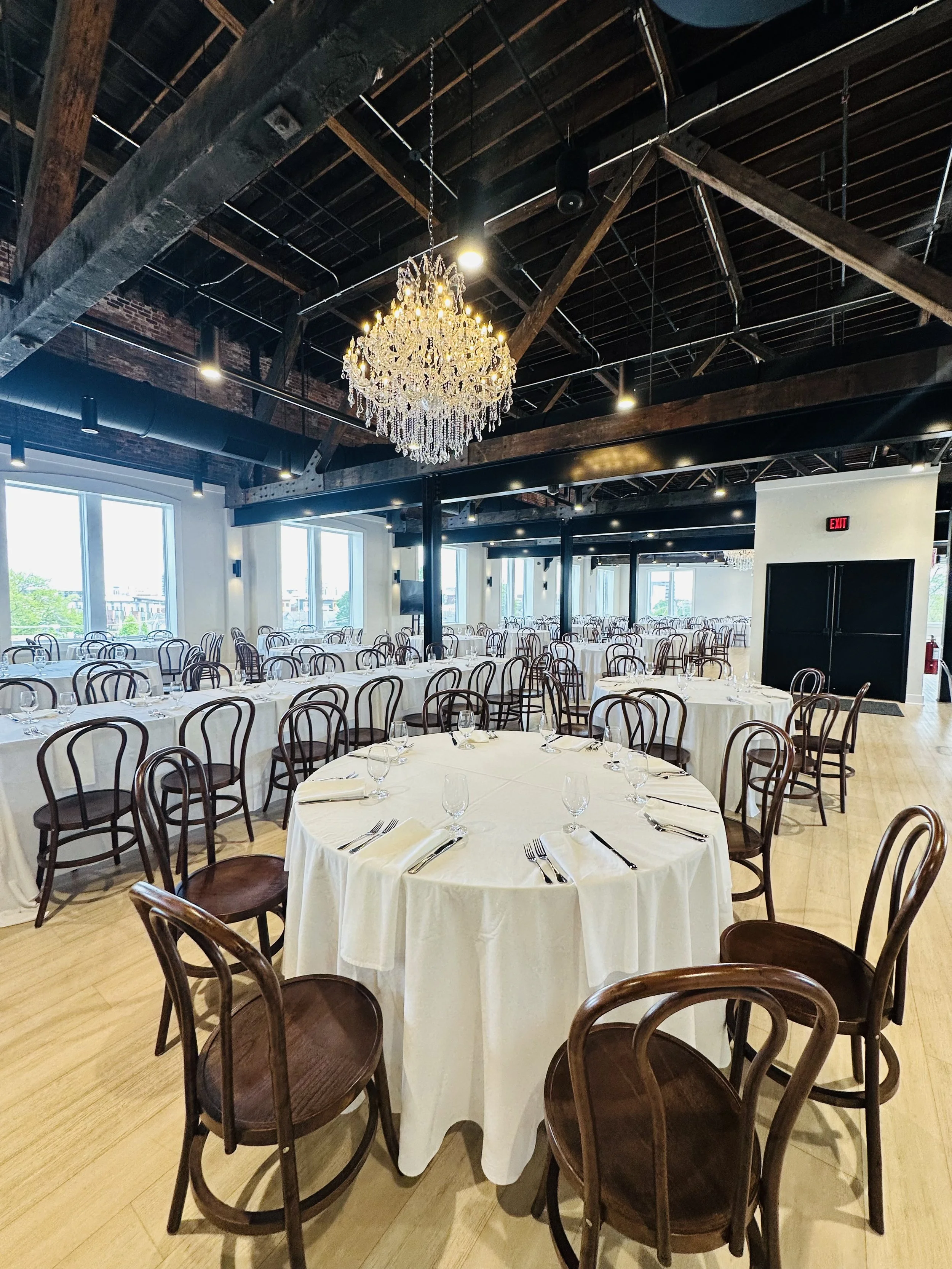 A large banquet hall with numerous tables covered with white tablecloths and set with silverware and glasses. Wooden chairs surround the tables. A chandelier hangs from the high ceiling with visible wooden beams. Large windows let in natural light, a