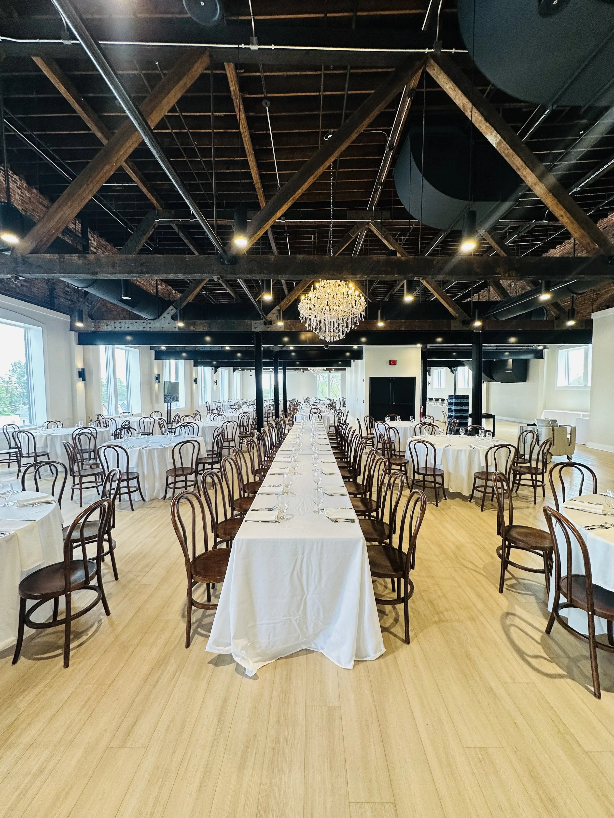 A banquet hall with long tables covered in white tablecloths and surrounded by wooden chairs. The hall has a high ceiling with exposed wooden beams, industrial-style ductwork, and a large chandelier hanging from the ceiling. Large windows allow natur