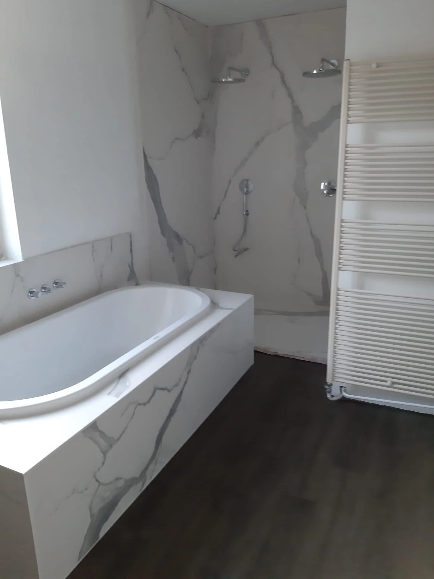 Badkamer met marmeren wand, een wit ingebouwd bad en handdoekradiator. Douche voor twee personen met twee douchekoppen in marmeren wand. Badkamerrenovatie SVO Projects in Zwijnaarde