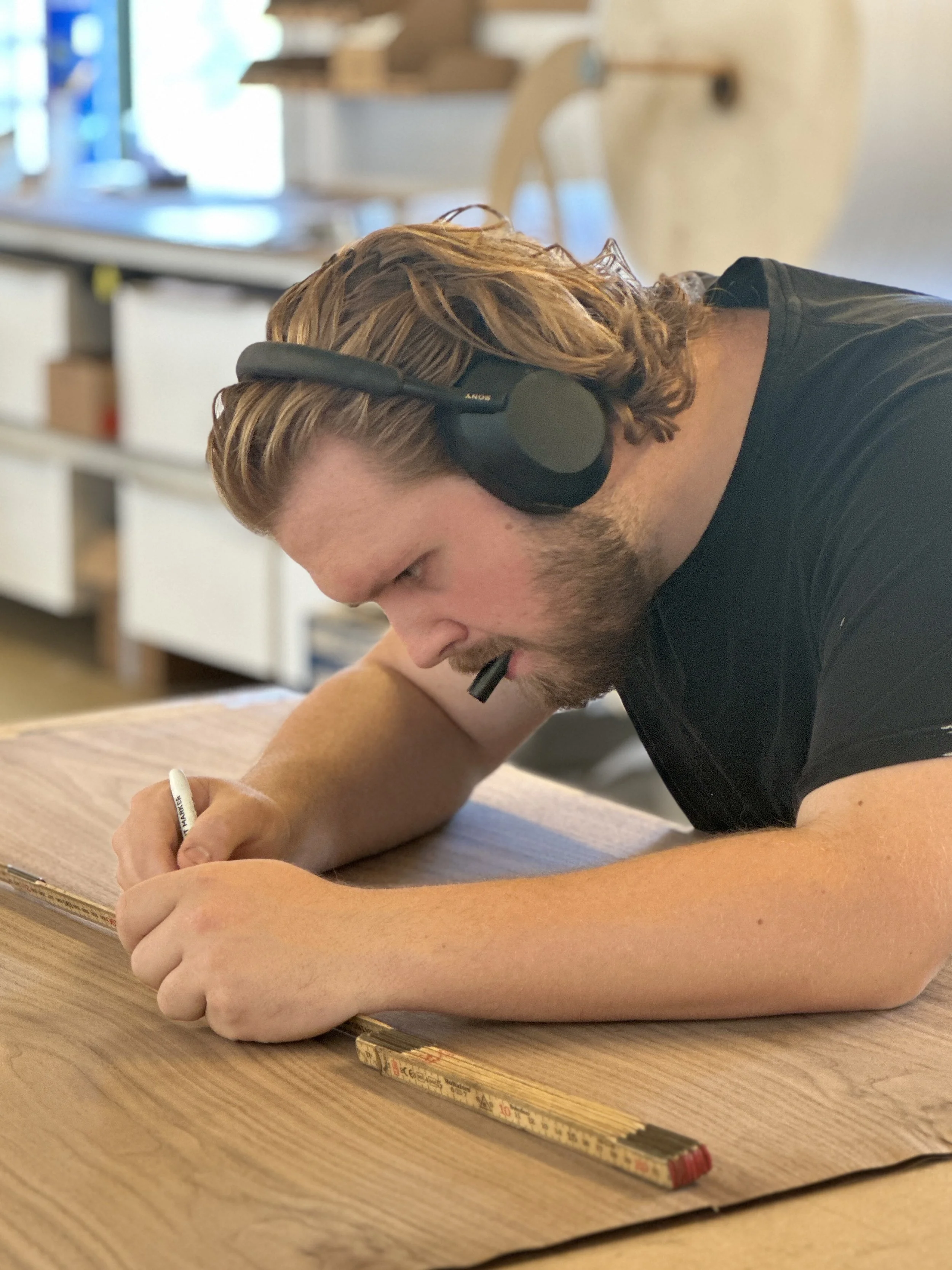 En mand med brunt, bølget hår og skæg, der arbejder i et værksted eller på et atelier, hvor han skriver på en skitse eller tegner på en træplade. Han bruger en sort pen og har hovedtelefoner på, mens han fokuserer på sit arbejde.