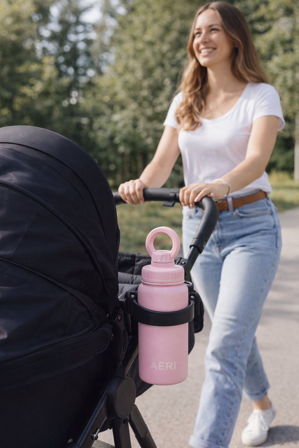 A woman pushing a stroller with a pink water bottle attached in an outdoor park setting.