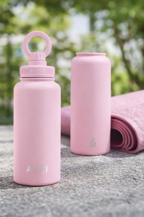 Pink water bottle with a lid and loop, next to a rolled pink yoga mat, outdoors with trees in the background.